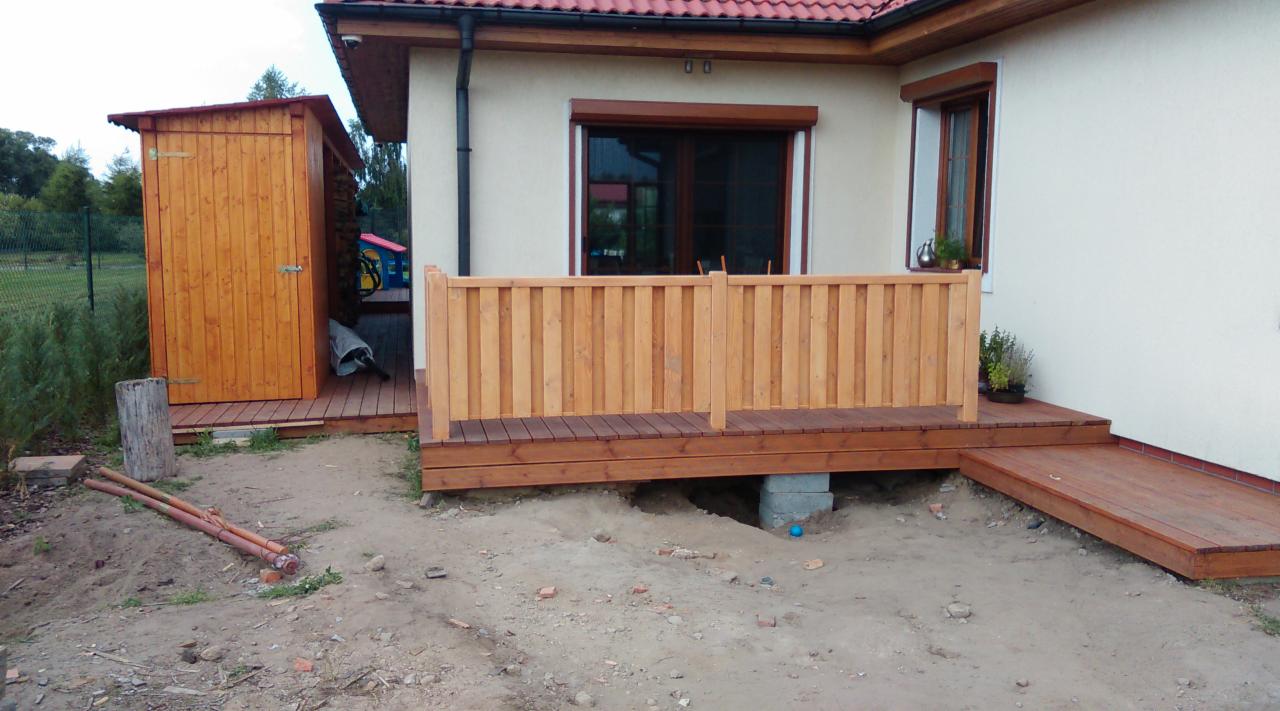 Drewniany taras z balustradą i schodami przy domu z beżową elewacją. Widoczny domek narzędziowy z drewna. Ujęcie z poziomu gruntu, z widocznymi elementami podkonstrukcji.