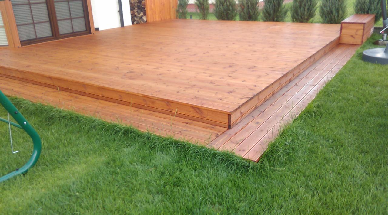 Drewniany taras z niskimi schodami na trawie, z widocznymi słojami drewna i subtelnym oświetleniem, przylegający do budynku z brązowymi oknami.