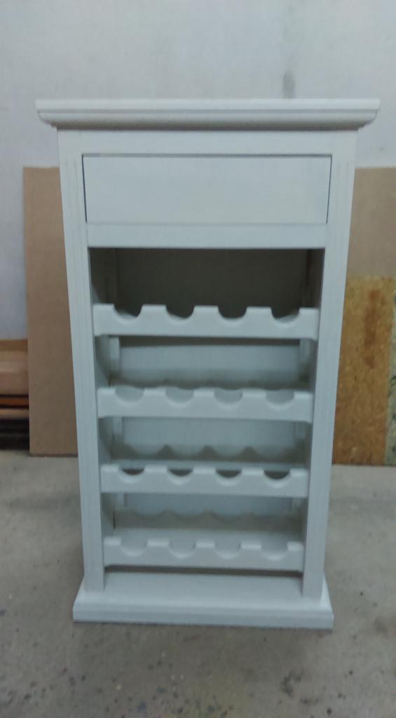 Biały, drewniany regał na wino z szufladą u góry, wykonany w stylu klasycznym. Widoczne półki na butelki i prosta forma mebla. Stoi na szarej podłodze.