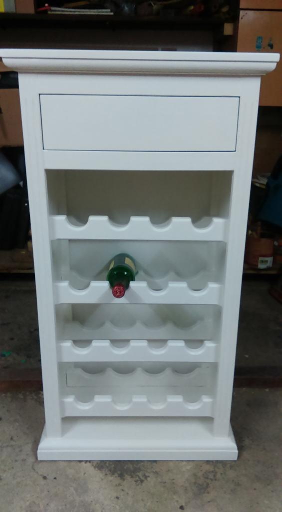 Biały, drewniany regał na wino z szufladą u góry, z jedną butelką wina na środkowej półce. Widoczny detal wykończenia górnej krawędzi. Całość w stylu rustykalnym.