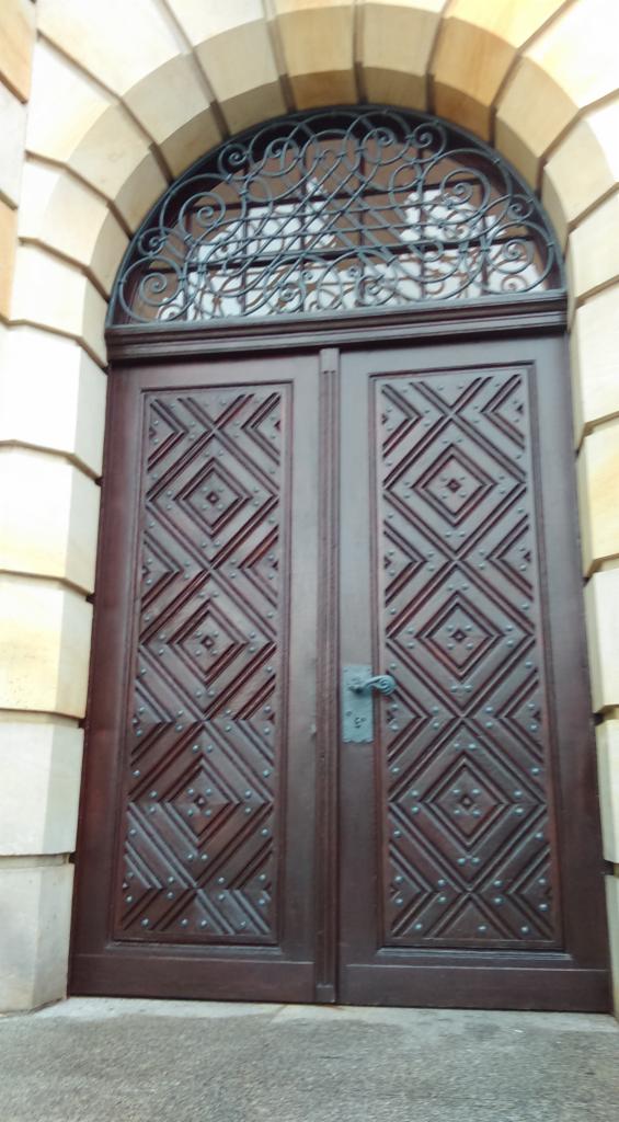 Dwuskrzydłowe, ciemne, drewniane drzwi z romboidalnymi zdobieniami i metalową kratą nadprożową, osadzone w kamiennym łuku.