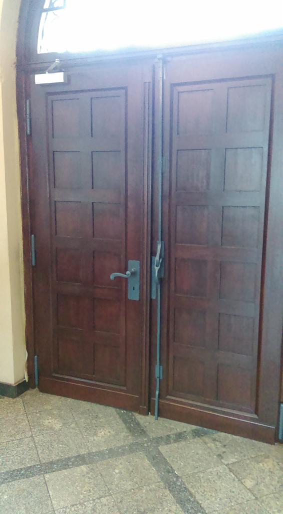 Ciemne, drewniane drzwi dwuskrzydłowe z metalowymi okuciami i szybą u góry, widoczne z bliska na tle podłogi wyłożonej płytkami. Zamek i klamka po lewej stronie.