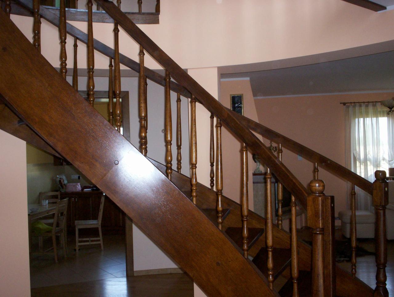 Drewniane schody z balustradą w odcieniach brązu, z widocznymi detalami tokarskimi na słupkach. W tle jasne ściany i widok na wnętrze domu.