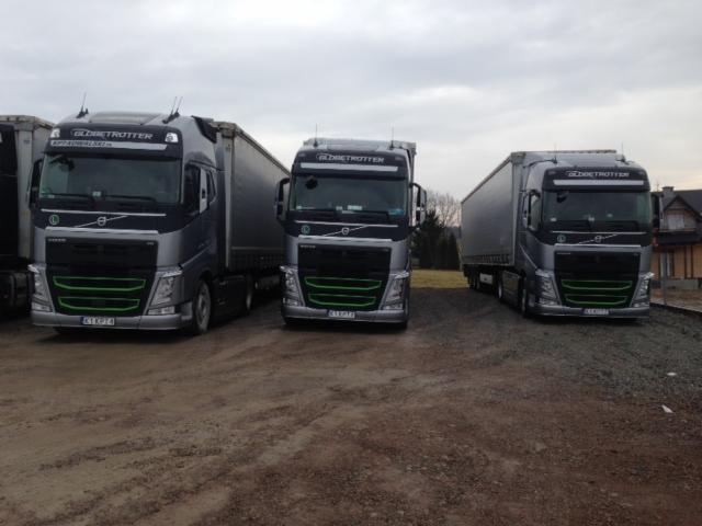 Trzy srebrne ciężarówki Volvo Globetrotter z zielonymi akcentami, ustawione w rzędzie na żwirowym placu. Jedna ciężarówka częściowo zasłonięta po lewej.