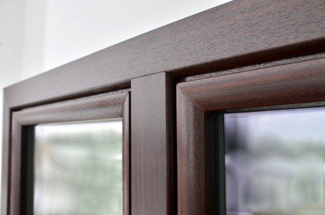 Zbliżenie na okno z brązową ramą o wyraźnej strukturze drewna. Widoczny fragment szyby i biała ściana w tle. Kąt ujęcia podkreśla detale wykończenia ramy.
