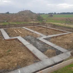 Zalane betonem ławy fundamentowe w wykopie, z widocznymi zbrojeniami i deskowaniem, na tle ziemi i pola uprawnego pod pochmurnym niebem.