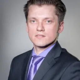 Portret mężczyzny w garniturze i fioletowym krawacie na szarym tle.