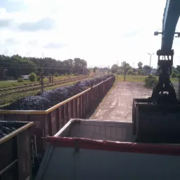 Kopalniany dźwig z łyżką ładuje ciemny materiał sypki z wagonów kolejowych do skrzyni ciężarówki na tle torów i zieleni.