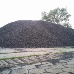 Góra czarnego, drobnego materiału na betonowym placu, z widocznymi płytami chodnikowymi i zielenią w tle.