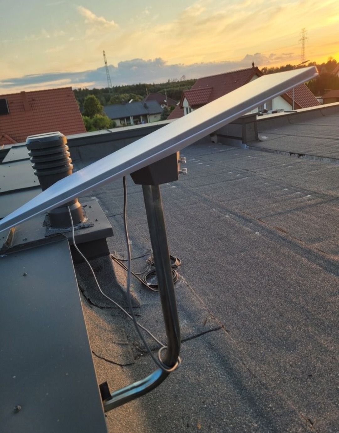 Antena satelitarna zamontowana na dachu budynku z widokiem na zachodzące słońce. Metalowa konstrukcja, kable. Tło: domy jednorodzinne i słup wysokiego napięcia.