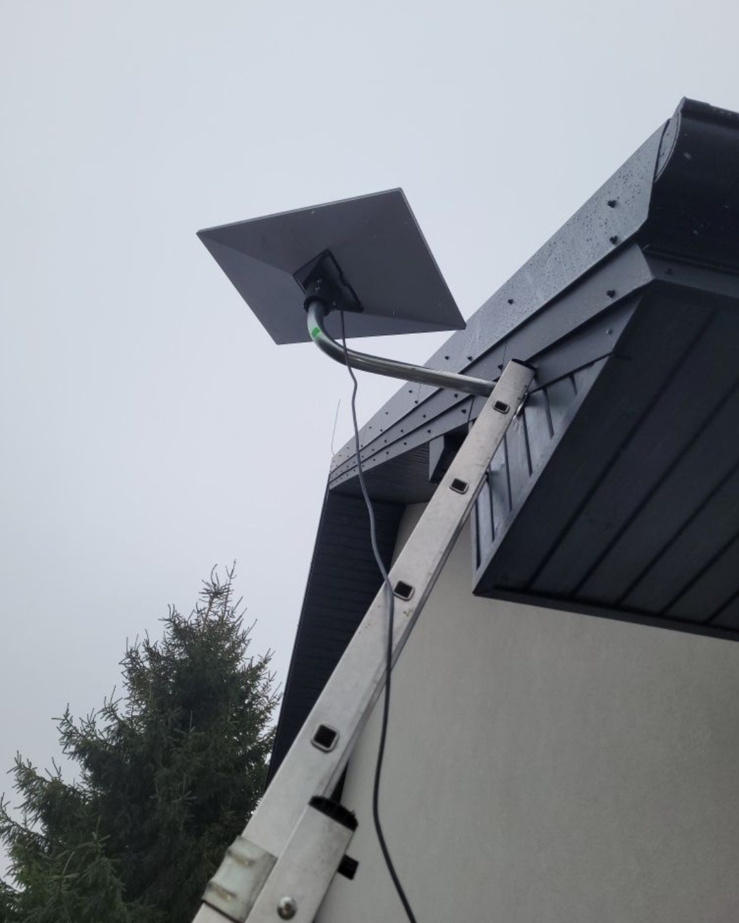 Antena satelitarna zamontowana na dachu domu w Bydgoszczy, widoczna drabina oparta o ścianę. Szara, pochmurna pogoda. Ujęcie z dołu.