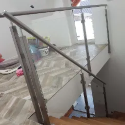 Szklana balustrada ze stalowymi słupkami i poręczą, zamontowana na schodach z drewnianymi stopniami i podłodze z płytek imitujących drewno, widok z góry.