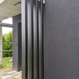 Nowoczesne, pionowe, czarne elementy metalowe tworzące ażurową ścianę przy wejściu do budynku z szarą, teksturowaną elewacją i kostką brukową na podłożu.
