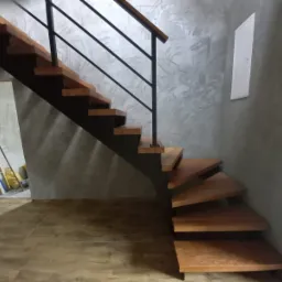 Drewniane schody z czarną metalową balustradą w nowoczesnym wnętrzu z szarą ścianą o dekoracyjnej fakturze i drewnianą podłogą.