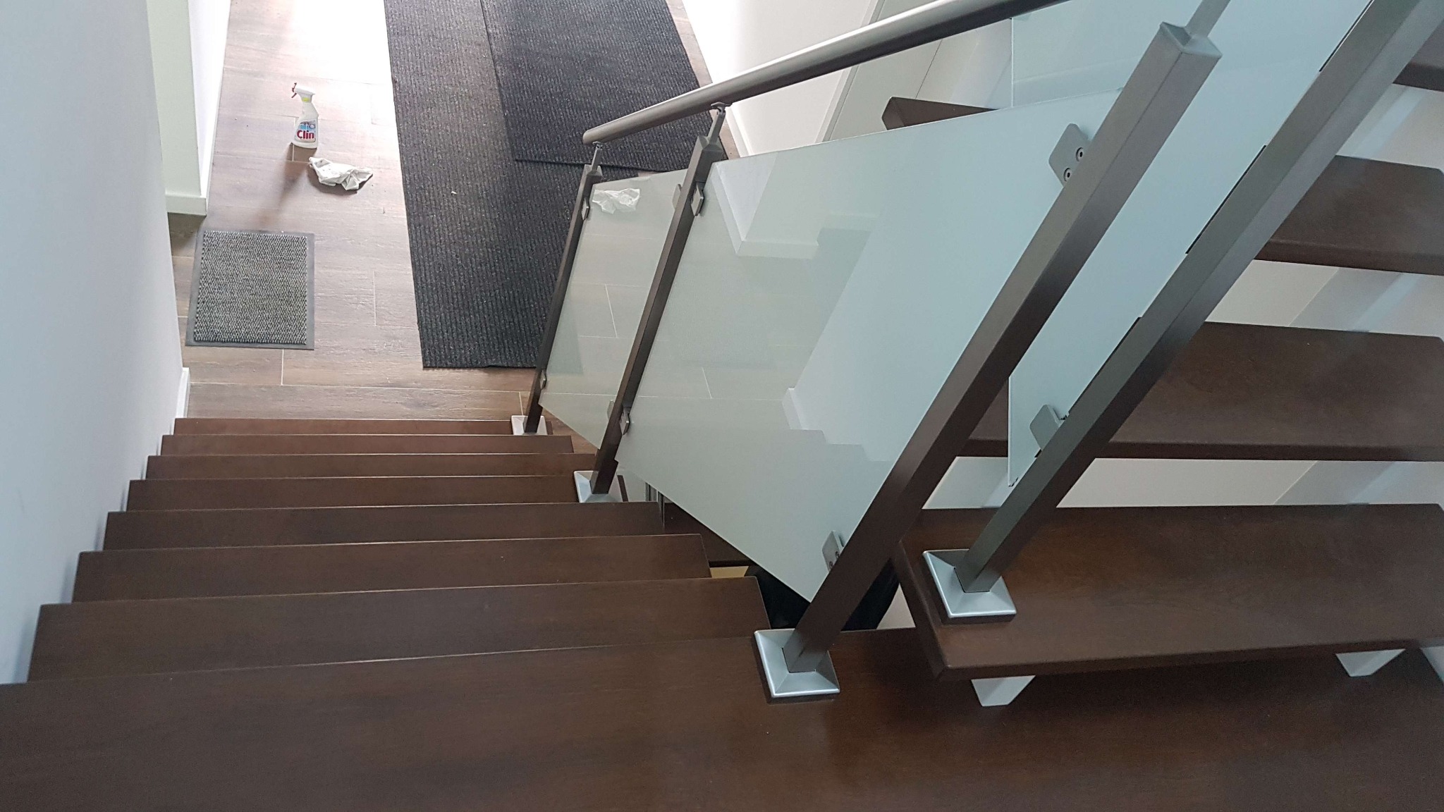Widok z góry na brązowe schody drewniane z balustradą ze szkła mlecznego i metalu, z widocznymi elementami mocującymi szkło do słupków, na podłodze widoczny dywanik i butelka środka czyszczącego.