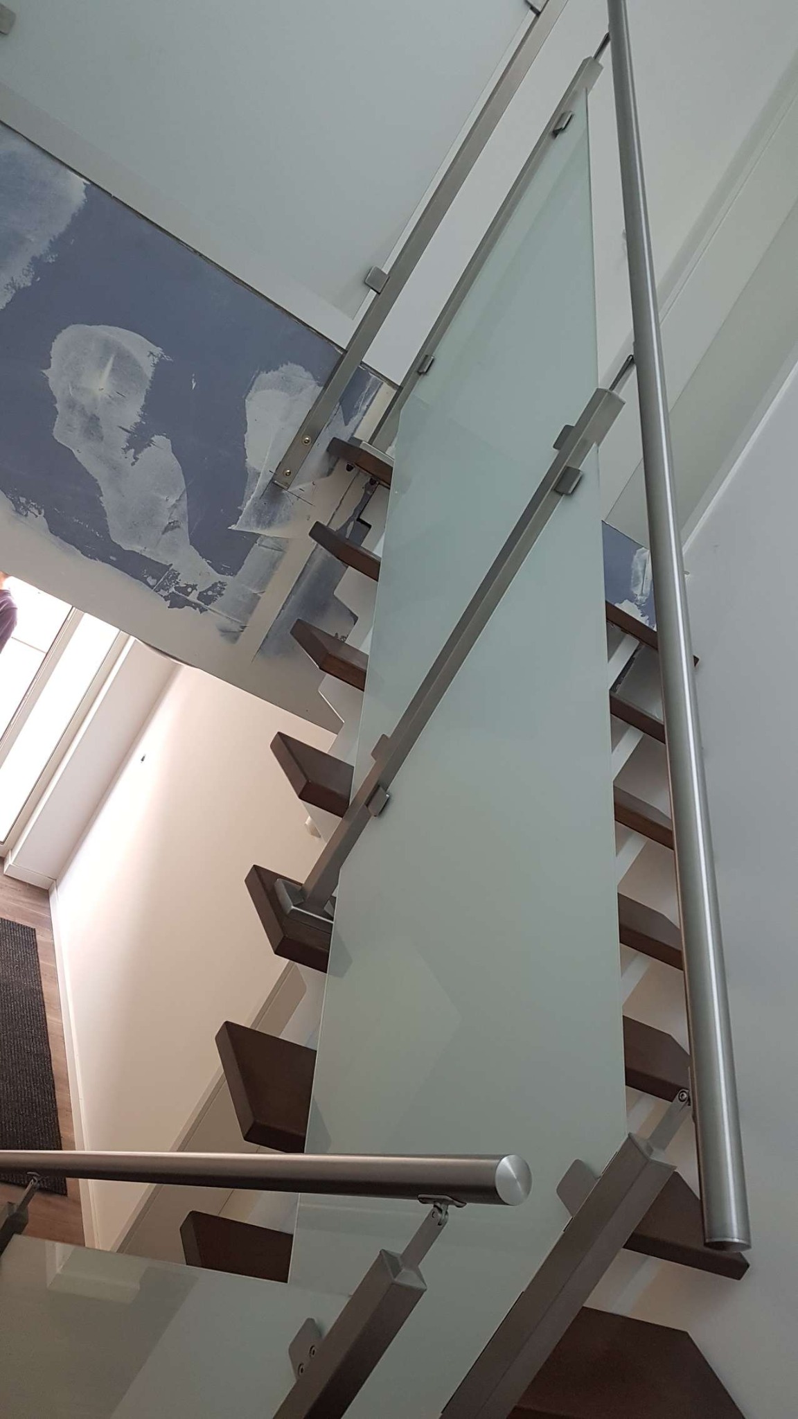 Nowoczesne schody z drewnianymi stopniami, balustradą ze szkła i metalowymi poręczami, widoczne ślady prac wykończeniowych na suficie.