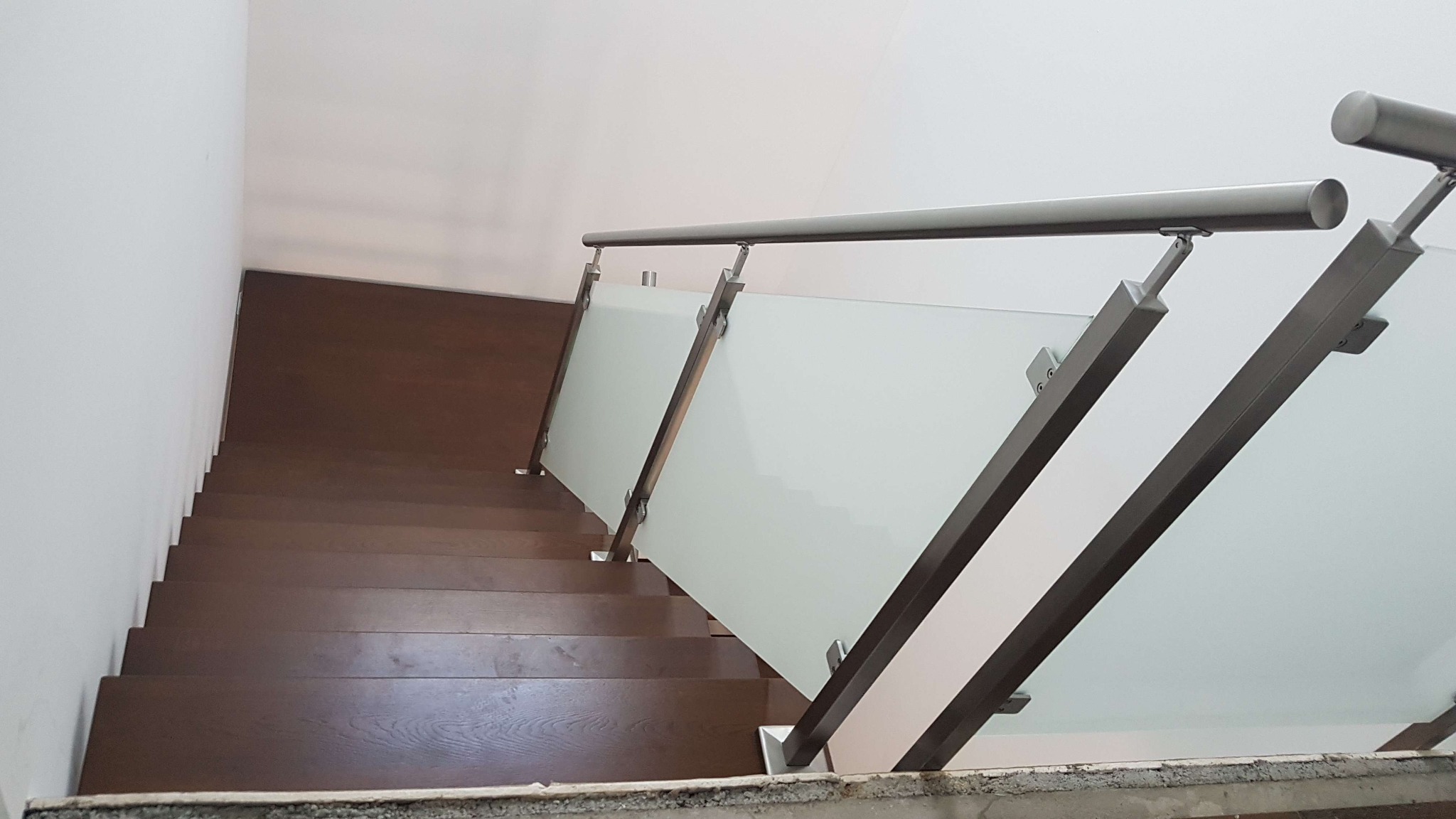 Widok z góry na drewniane schody z balustradą ze szkła matowego i metalowymi poręczami, montowanymi na metalowych słupkach.