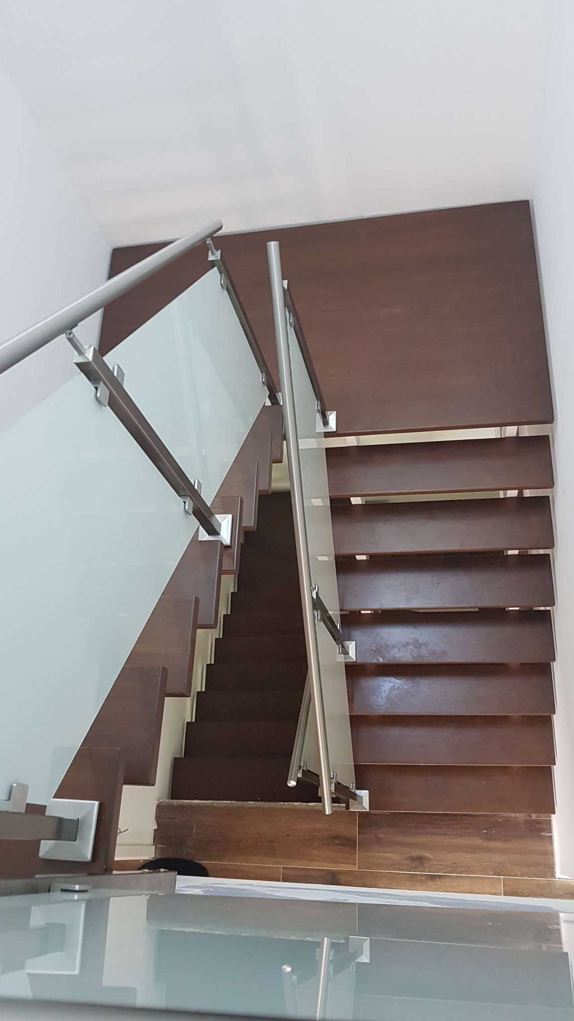 Klatka schodowa z drewnianymi stopniami, szklaną balustradą i metalowymi poręczami, widok z góry.