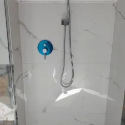 Białe płytki imitujące marmur w kabinie prysznicowej z chromowaną baterią i słuchawką prysznicową, widoczne łączenia płytek.