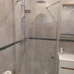 REMBUD Usługi Remontowo-Budowlane - Kabina prysznicowa z deszczownicą i baterią termostatyczną, wykończona płytkami imitującymi marmur z dekoracyjnym pasem mozaiki, widoczna umywalka z szafką w tle.