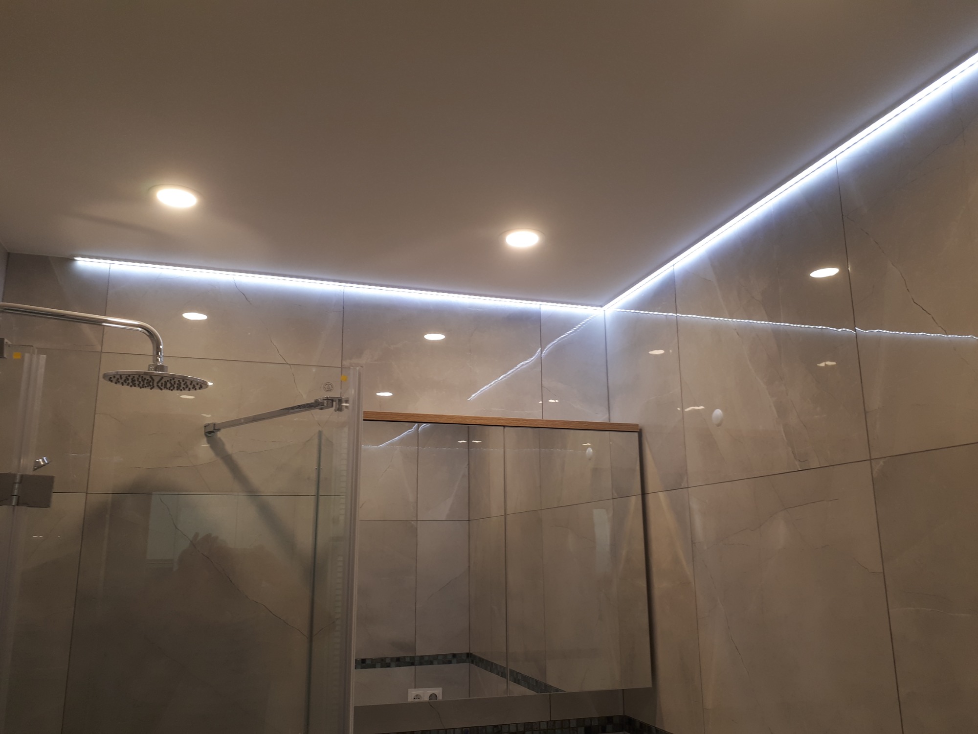 Wykończona łazienka z kabiną prysznicową typu walk-in, oświetleniem LED podsufitowym i płytkami imitującymi marmur na ścianach.