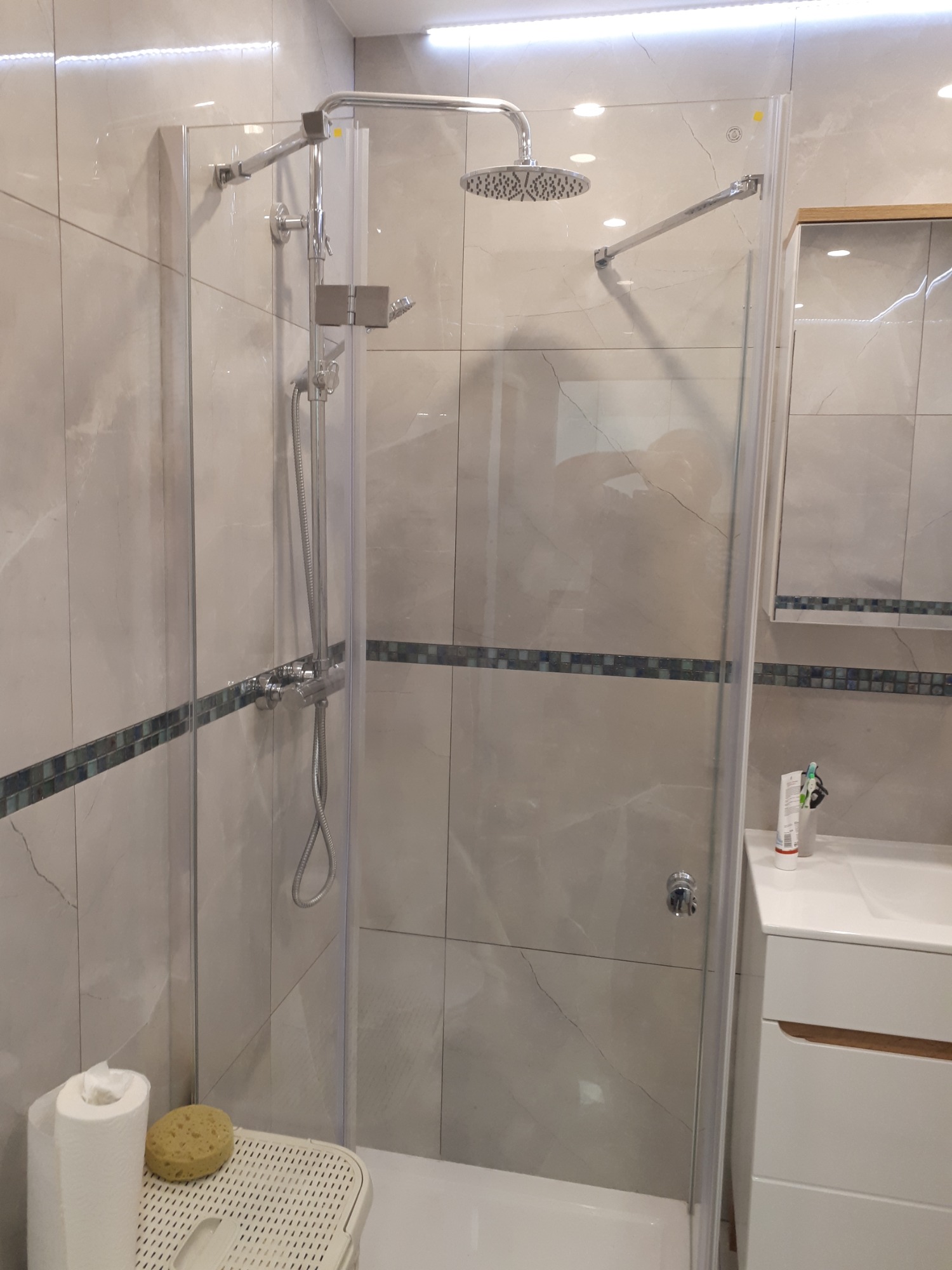 Kabina prysznicowa z deszczownicą i baterią termostatyczną, wykończona płytkami imitującymi marmur z dekoracyjnym pasem mozaiki, widoczna umywalka z szafką w tle.