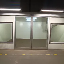 Przesuwne, szklane drzwi do chłodni z dwoma okienkami po bokach, widoczne oświetlenie sufitowe i żółte oznaczenia na podłodze.