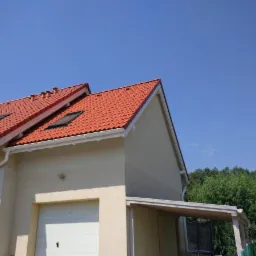 Nowoczesny dom z czerwonym dachem, oknami dachowymi i białymi rynnami w słoneczny dzień.