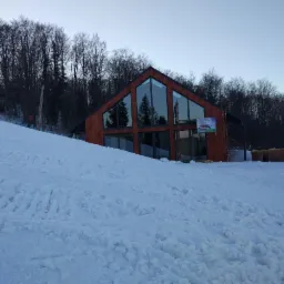 Nowoczesny dom z drewna z dużymi przeszkleniami położony na stoku narciarskim, częściowo zasypany śniegiem, widoczny baner reklamowy.
