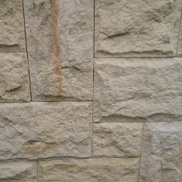 Szczegółowy widok elewacji z piaskowca w odcieniach beżu, z widocznymi nieregularnymi kształtami kamieni i fugami, z subtelnym zaciekem na jednym z elementów.