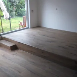 Ułożona podłoga z płytek imitujących drewno, z widocznym stopniem i fragmentem zabudowy okna. Na zewnątrz widok na ogród.