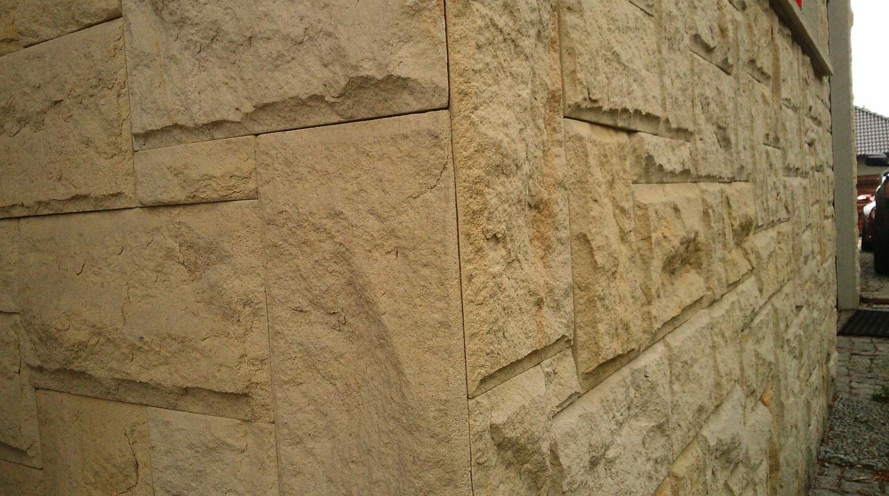 Elewacja budynku wykończona płytami z piaskowca w odcieniach beżu, widok na narożnik budynku z bliska, z widocznym fragmentem ogrodzenia i samochodu w tle.