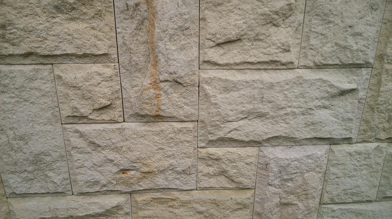 Szczegółowy widok elewacji z piaskowca w odcieniach beżu, z widocznymi nieregularnymi kształtami kamieni i fugami, z subtelnym zaciekem na jednym z elementów.