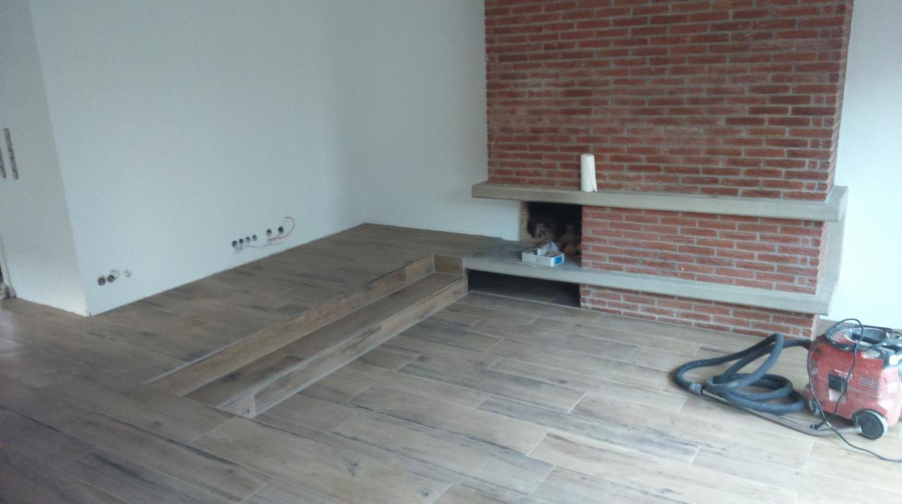 Ukończona podłoga z płytek imitujących drewno z dwustopniowym podwyższeniem, widoczny murowany kominek z betonowymi półkami i odkurzacz przemysłowy w rogu pokoju.