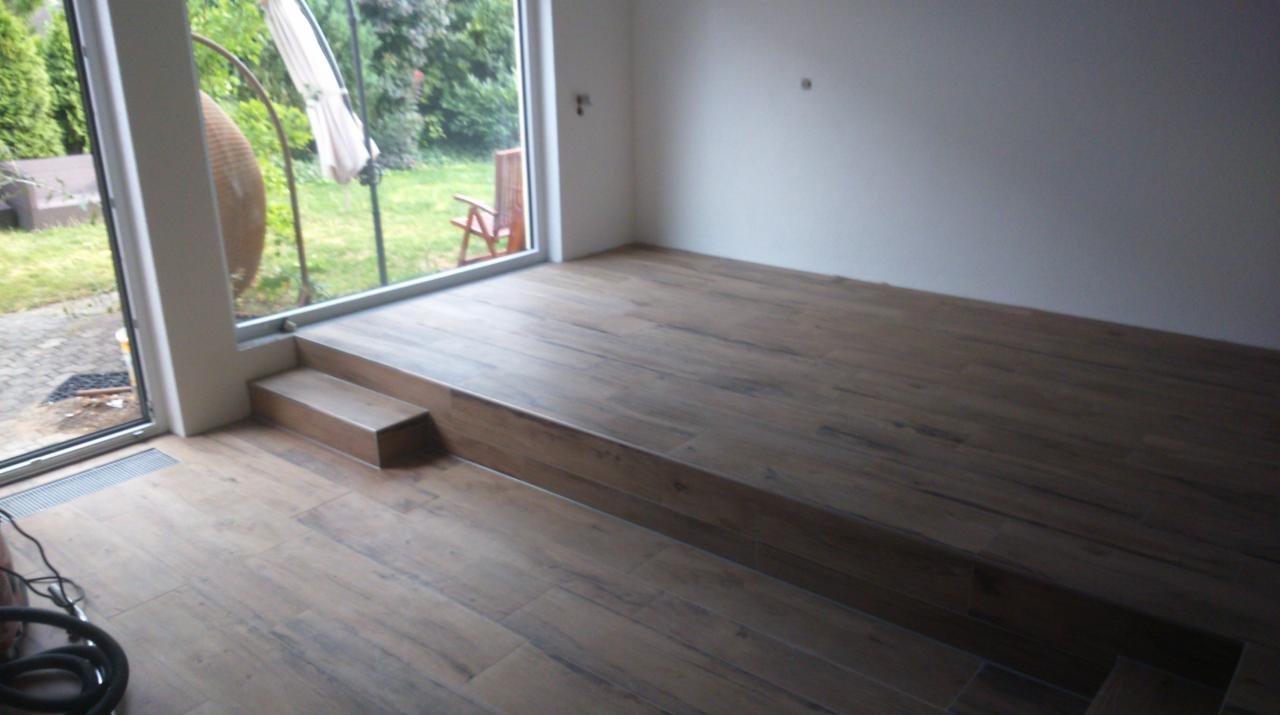 Ułożona podłoga z płytek imitujących drewno, z widocznym stopniem i fragmentem zabudowy okna. Na zewnątrz widok na ogród.