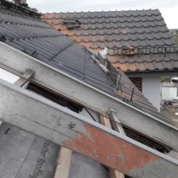 Widok z góry na konstrukcję dachu w trakcie budowy, z widoczną membraną dachową, ociepleniem styropianowym i częściowo ułożoną dachówką ceramiczną, a w tle nowo budowane budynki mieszkalne.