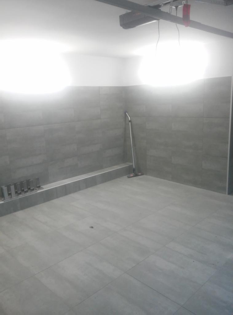 Wykończona łazienka z szarymi płytkami podłogowymi i ściennymi, widoczna listwa przypodłogowa i narzędzia do układania płytek oraz mop przy ścianie.