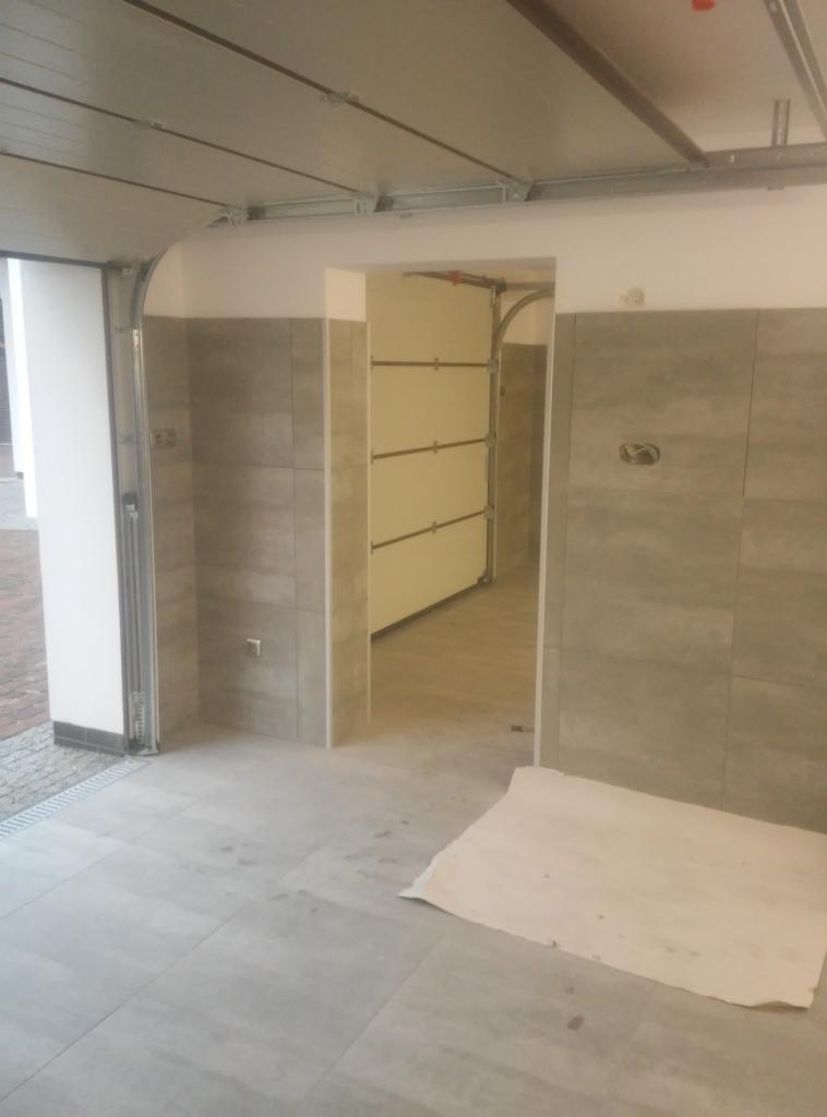 Wykończone wnętrze garażu z szarą glazurą na ścianach i podłodze, widoczne otwarte drzwi garażowe i zabezpieczająca folia na podłodze.