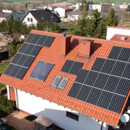 Instalacja paneli słonecznych na dachu domu z czerwoną dachówką, widok z góry na tle zabudowań i drzew