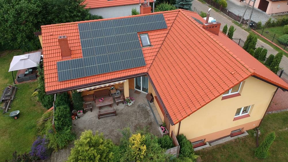 Dom jednorodzinny z pomarańczową dachówką i panelami fotowoltaicznymi na dachu, widok z góry, patio z meblami ogrodowymi.