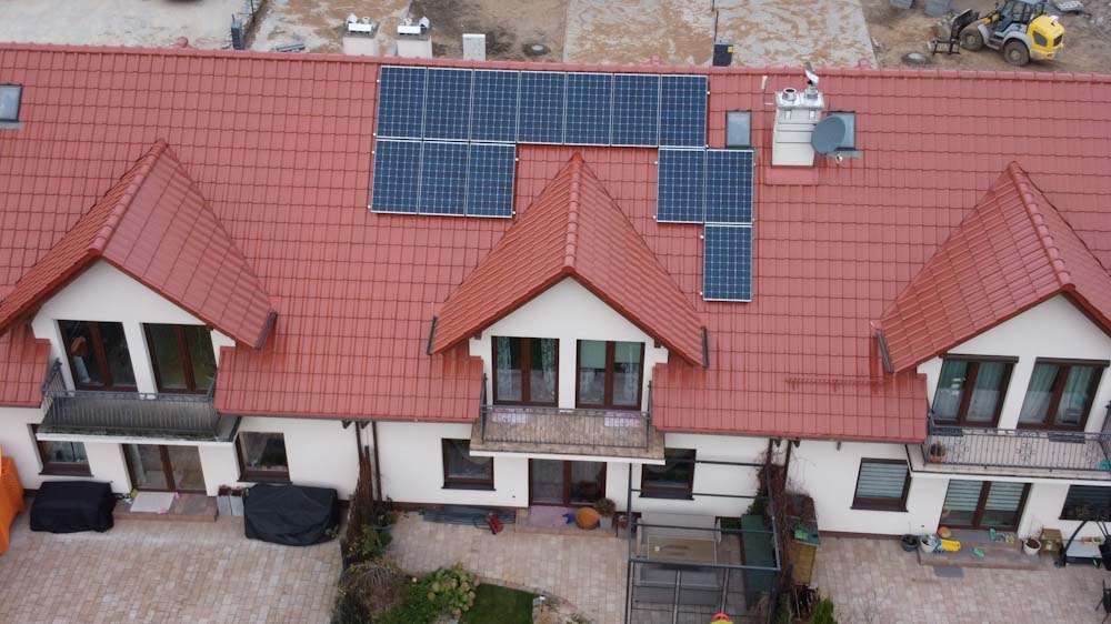Widok z góry na szeregowy dom jednorodzinny z czerwoną dachówką i zainstalowanymi panelami słonecznymi; widoczny fragment placu budowy w tle.