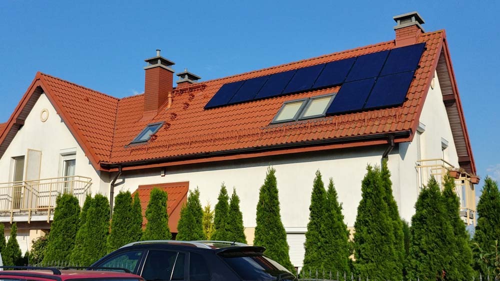 Dom jednorodzinny z czerwoną dachówką i panelami słonecznymi na dachu, widoczne okna dachowe, kominy i iglaki w ogrodzie.