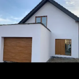 Nowoczesny, biały dom jednorodzinny z brązową bramą garażową, oknami i drzwiami, widoczny z zewnątrz w słoneczny dzień.