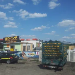 Budynek IZO-TAR z reklamą materiałów budowlanych, zaparkowany samochód firmowy, ciężarówka z plandeką z logo firmy, brama wjazdowa i błękitne niebo.