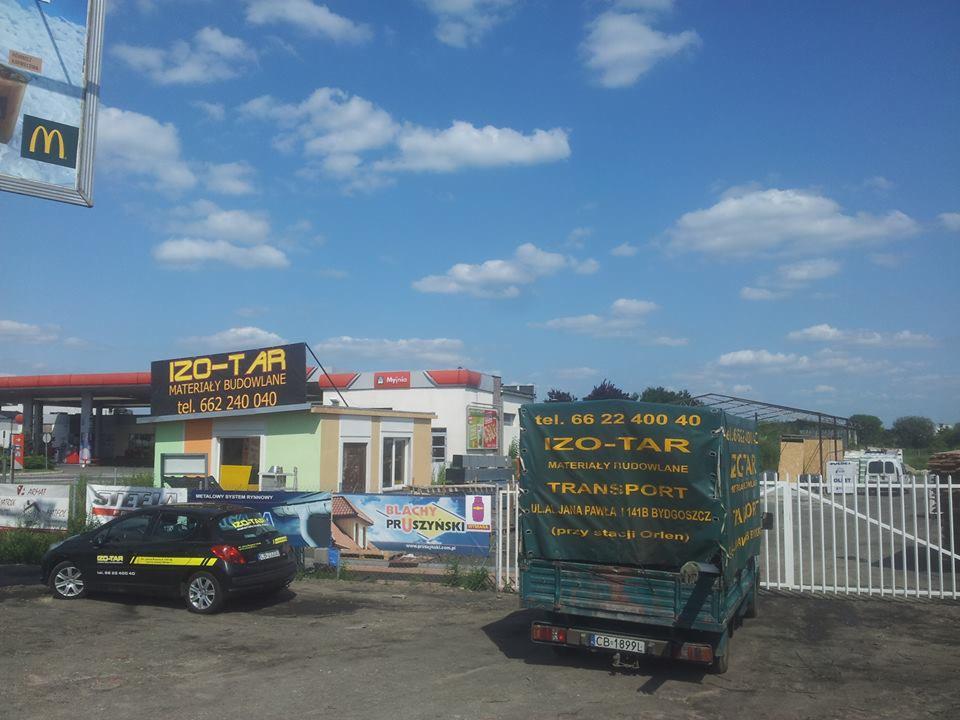 Budynek IZO-TAR z reklamą materiałów budowlanych, zaparkowany samochód firmowy, ciężarówka z plandeką z logo firmy, brama wjazdowa i błękitne niebo.