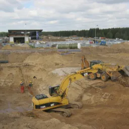 Żółta koparka CAT 320D w trakcie prac ziemnych przy głębokim wykopie, w tle budynek obsługi i teren budowy z hałdami piachu.