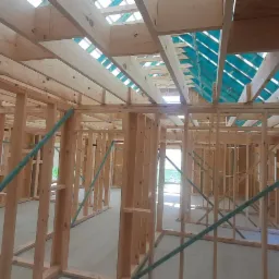 Surowa konstrukcja drewniana domu jednorodzinnego w trakcie budowy, widoczne ściany szkieletowe, strop z izolacją i otwory okienne.