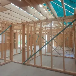 Wnętrze domu w trakcie budowy: drewniana konstrukcja szkieletowa ścian i dachu, widoczne płyty OSB, zabezpieczenie folią na dachu, zielone elementy mocujące.