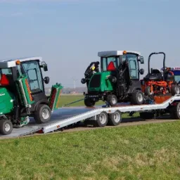 Niebieska laweta z najazdami przewozi dwa zielone traktorki do koszenia trawy i pomarańczowy wózek golfowy na tle zielonego pola i błękitnego nieba.
