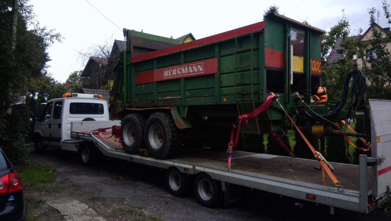 Przyczepa rolnicza Bergmann zabezpieczona pasami transportowymi na platformie lawety, widok z boku, w tle zabudowa jednorodzinna.